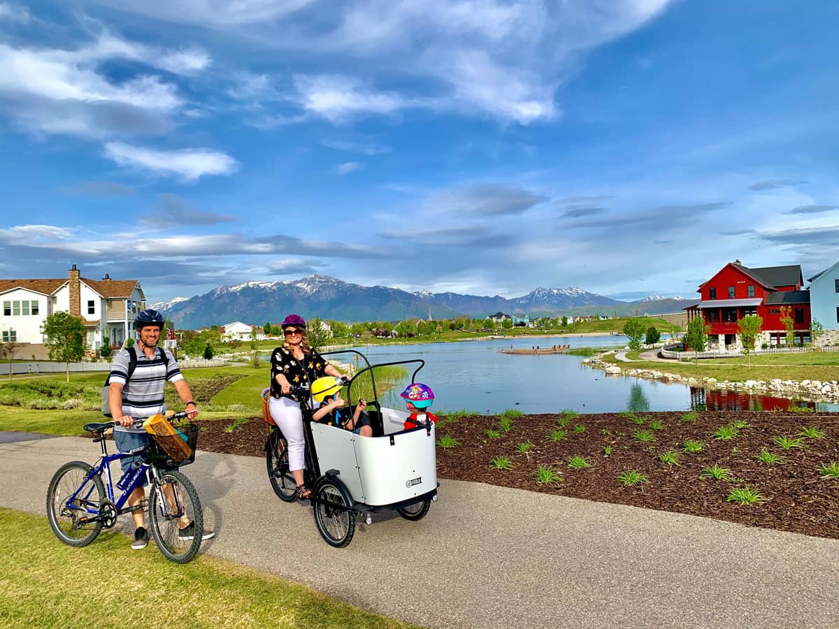 A family enjoying a bike ride with a cargo bike against a scenic backdrop with mountains and a lake.